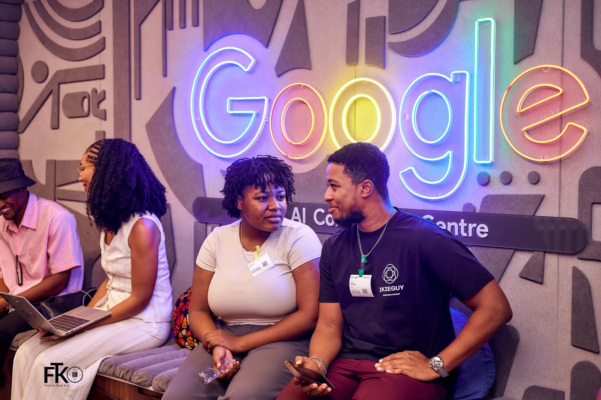 Two attendees in conversation on the bench under the Google neon, one in an Ikieguy shirt